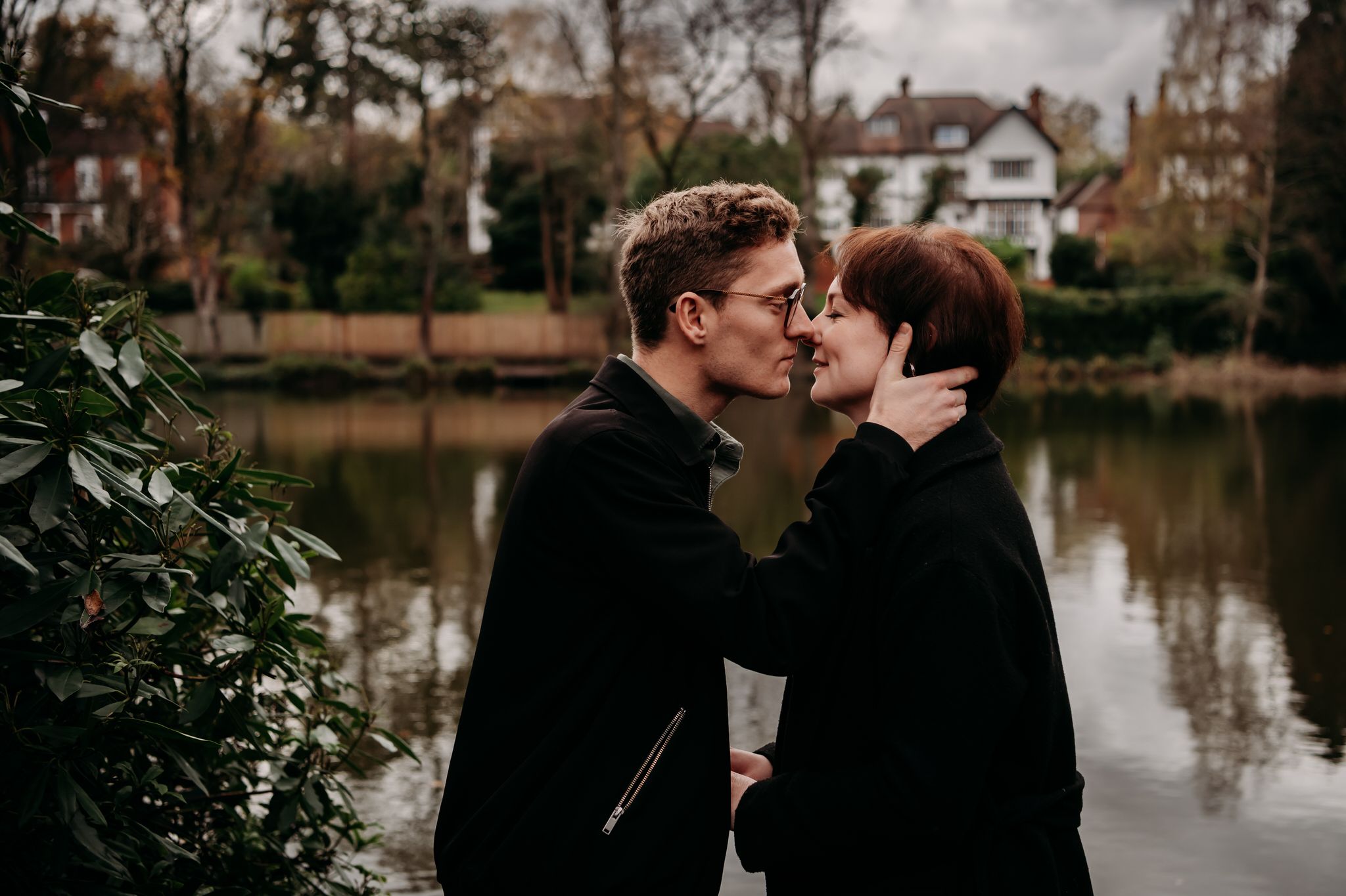 Couple embraces near a serene lake with fall foliage and houses in the background.