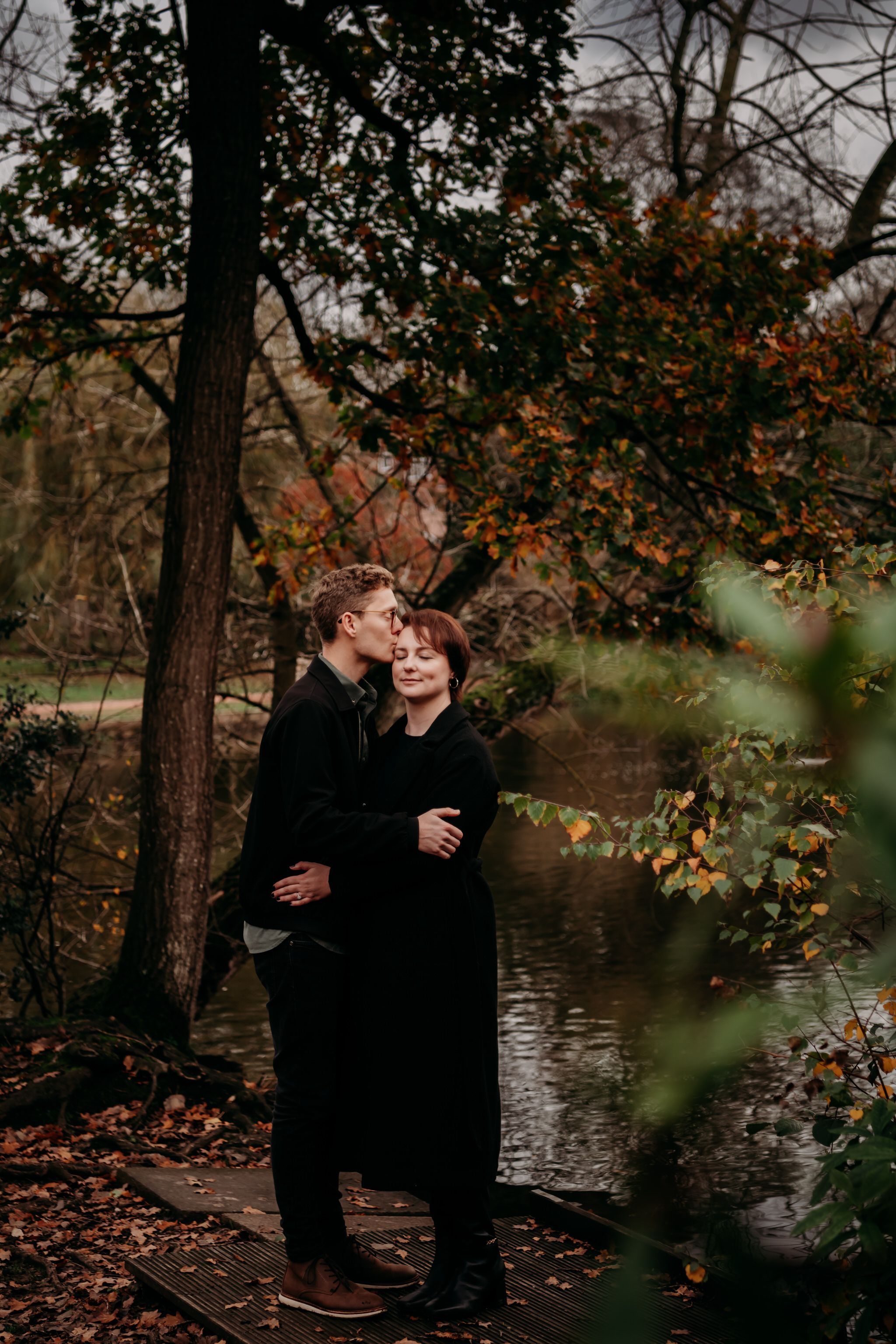 Couple sharing an intimate moment near a lake surrounded by fall foliage and trees.
