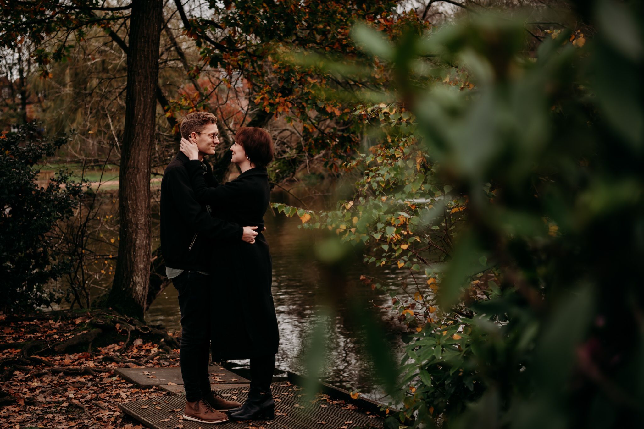 A romantic autumn scene by a pond with two people in black coats embracing among fallen leaves and trees.