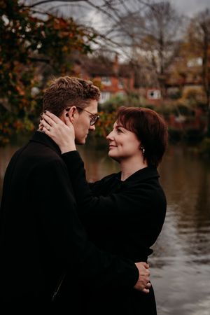 Two people in black coats share an intimate moment beside a lake during autumn, with red brick buildings in background.