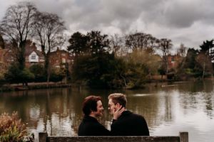 Moody engagement photos captured by a lake with dramatic cloudy skies and bare trees.