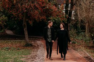 A couple walks together on a tree-lined autumn path with fallen leaves and warm lighting.