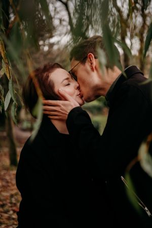 Romantic couple sharing an intimate kiss in a wooded area with autumn trees in the background.