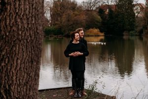 A romantic lakeside scene with a couple embracing among fall foliage and tree trunks.
