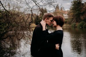 Tender moment captured between two people in black clothing against a moody lakeside backdrop.