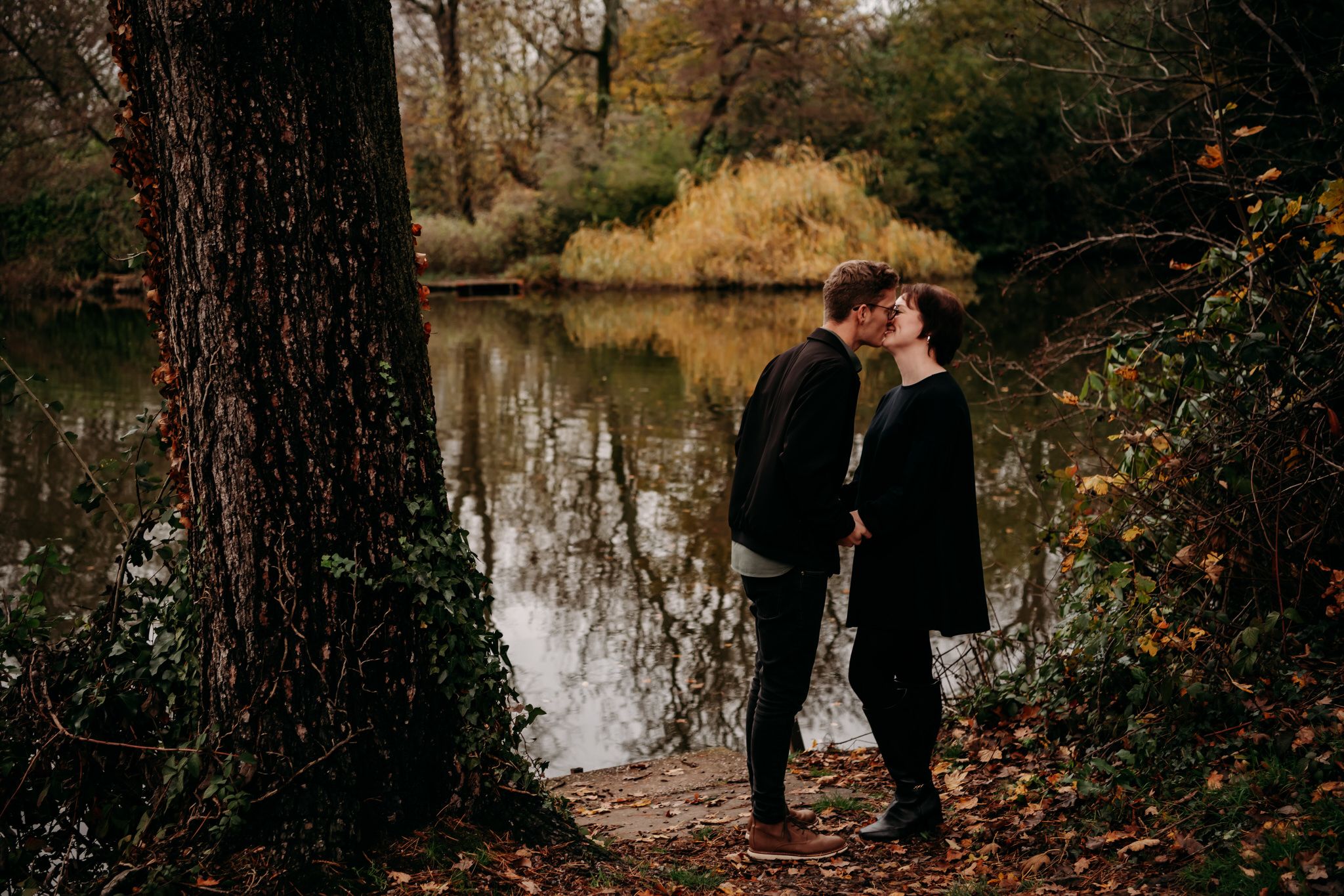 A couple shares an intimate moment near a lake surrounded by autumn trees and fallen leaves.