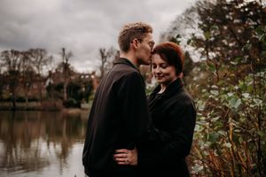 Two people share an intimate moment on a path near a tranquil lake surrounded by autumn foliage.