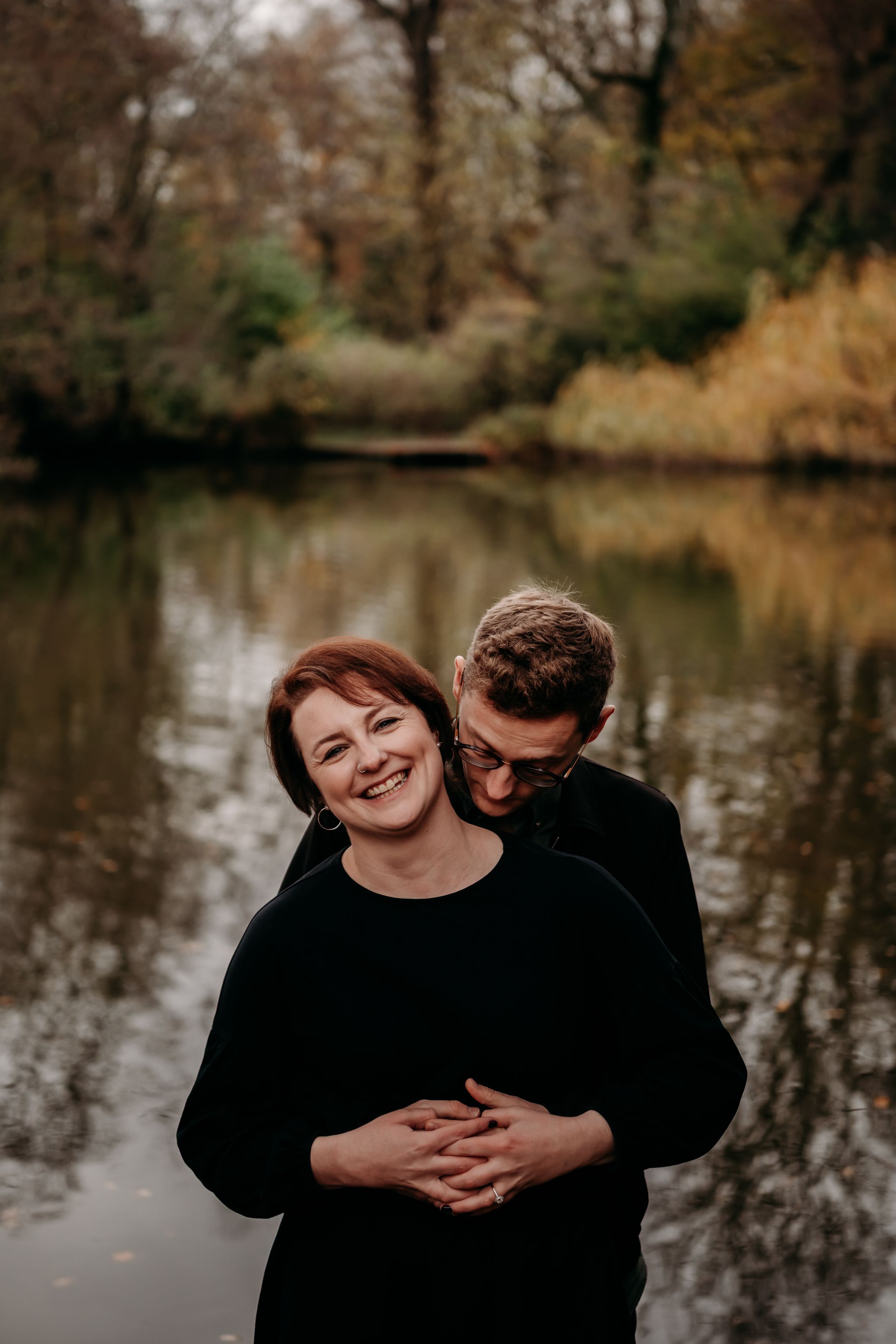 A romantic couple embraces by a tranquil autumn lake with bare trees reflected in the calm water.