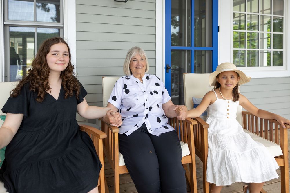People sit on wooden rocking chairs on a front porch of a white house with blue trim.
