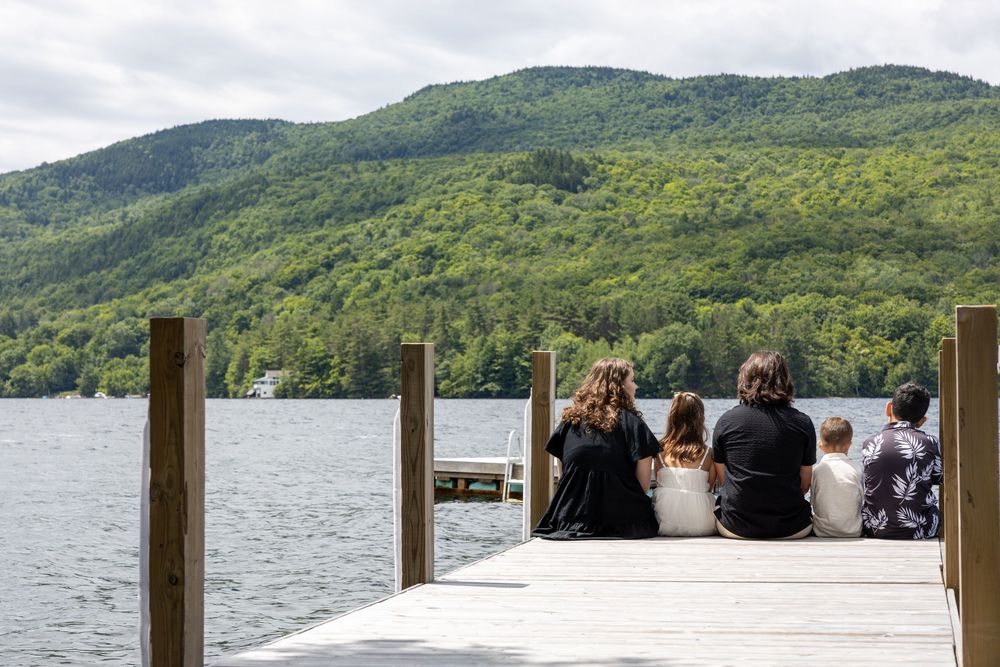 People sit on a wooden dock overlooking a serene lake surrounded by lush green mountains on a summer day.