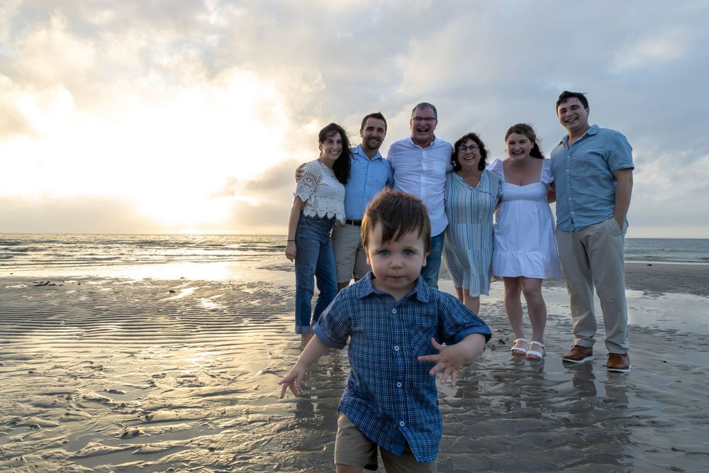 A joyful group stands on a beach at sunset while a child in blue runs playfully in the foreground.