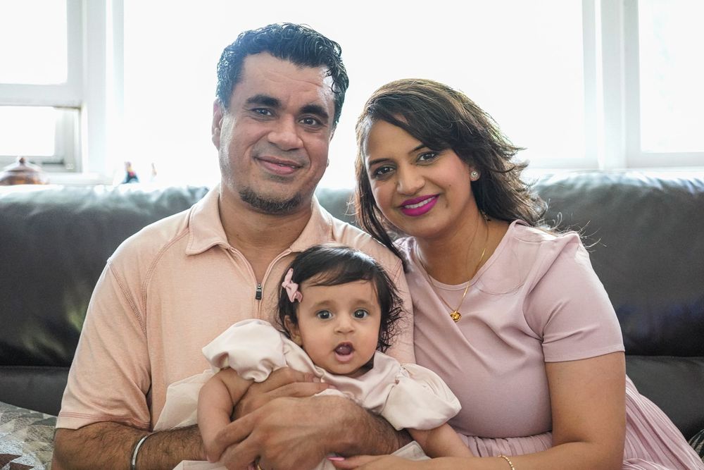 A family of three sits together on a grey couch, with the baby wearing white and parents in pink and peach tones.