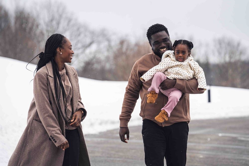 A family walks together on a snowy winter day, with one person holding a small child in a pink outfit.
