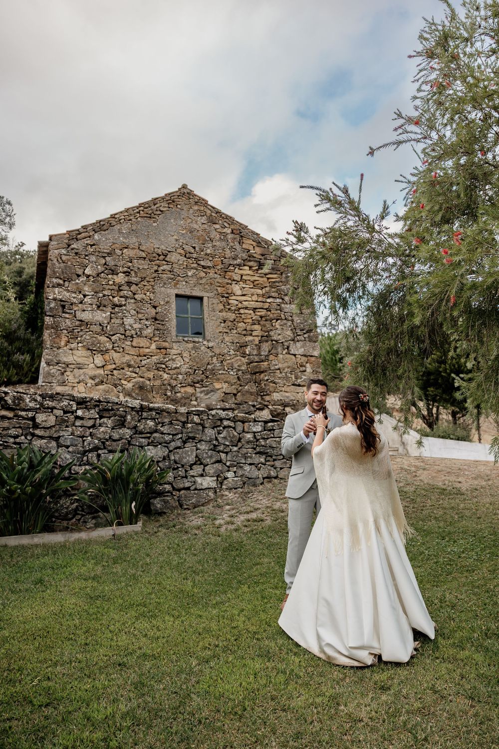 Bride and groom dancing outdoors on green grass in front of a rustic stone building on their wedding day.