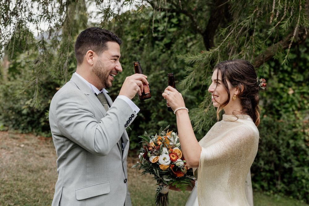 Bride and groom toasting with beer bottles outdoors, laughing joyfully on their wedding day.