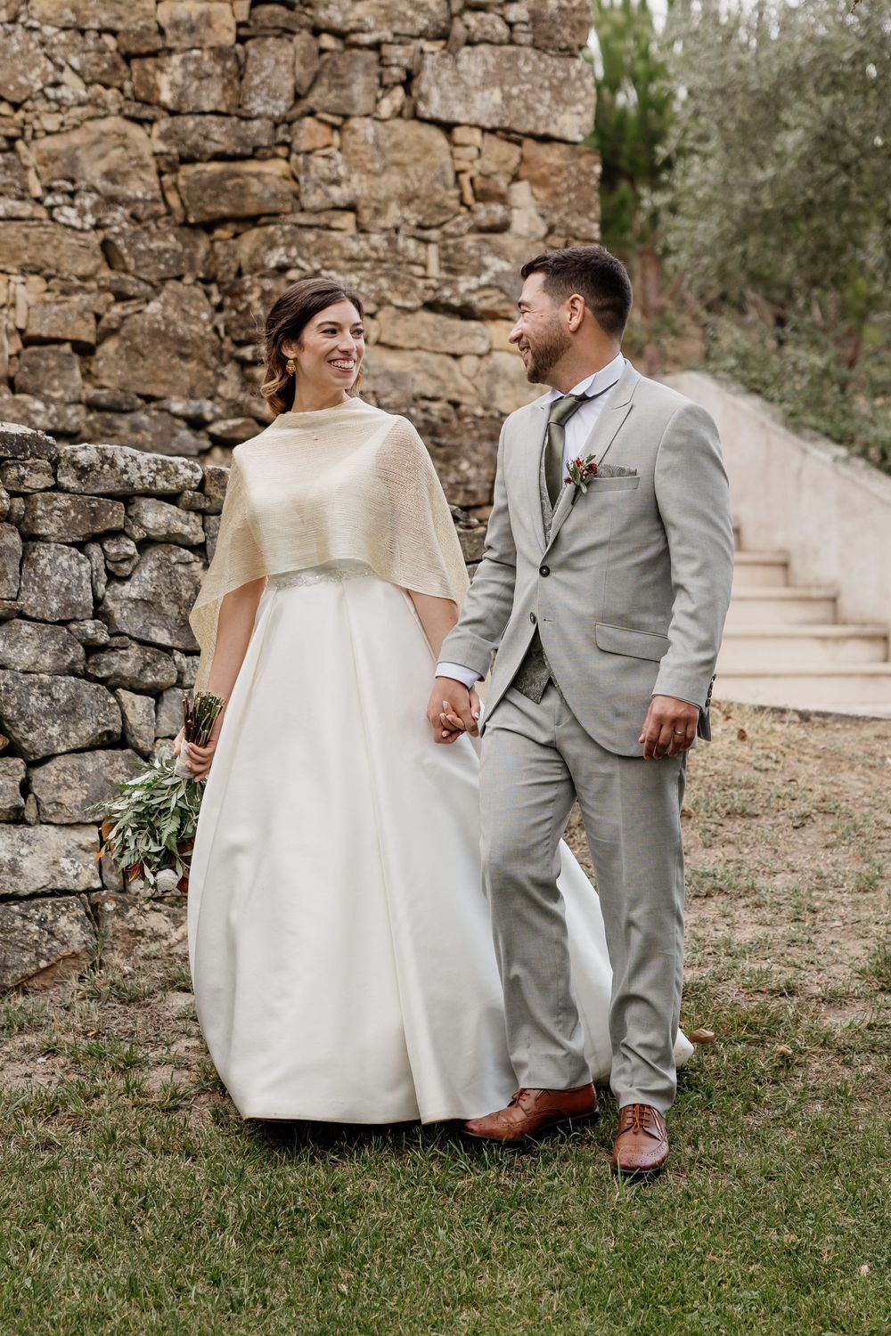 A bride and groom walk hand in hand, smiling at each other, beside a rustic stone wall on their wedding day.