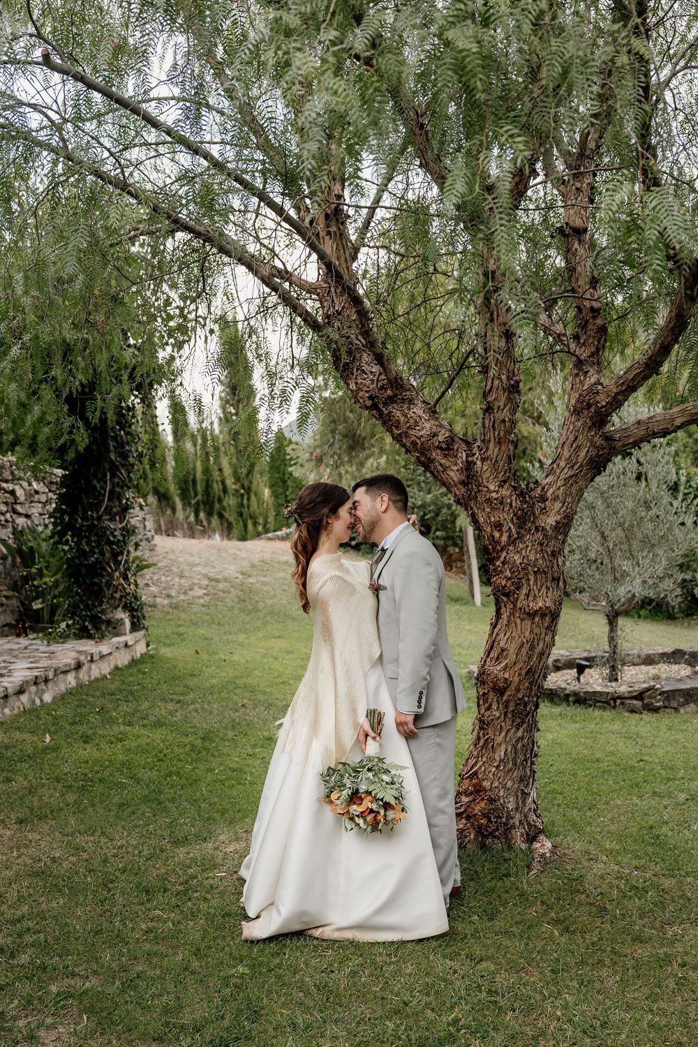 Bride and groom kissing under a tree in a lush garden, bride holding a colorful bouquet in a white gown.