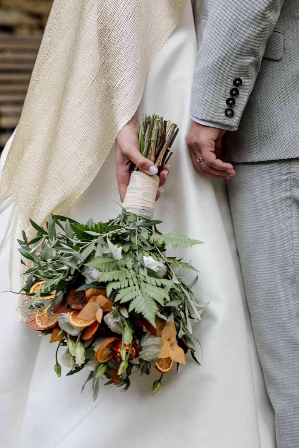 Bride holding a rustic wedding bouquet with greenery, ferns, orange slices, and flowers beside groom in gray suit.