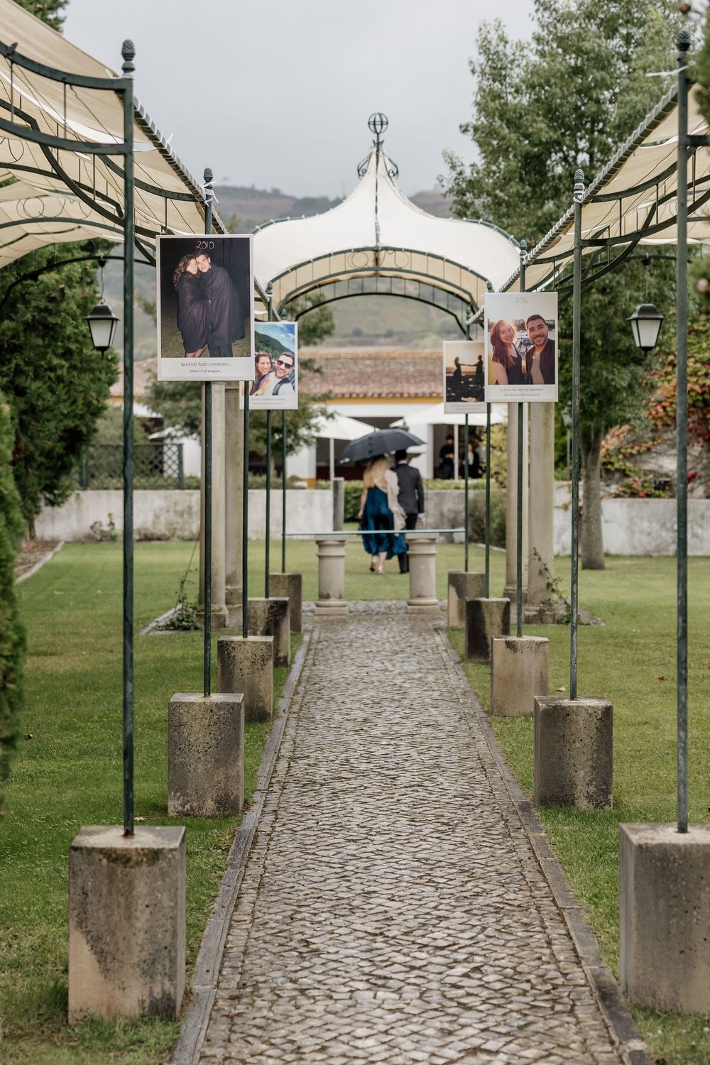 Cobblestone garden pathway lined with metal arches displaying photos, leading to a white tent pavilion on a rainy day.