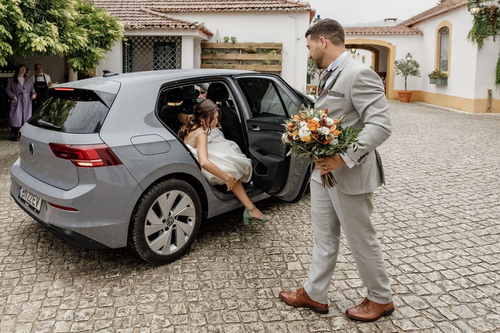 Groom in gray suit holds bouquet as bride exits gray Volkswagen Golf at elegant white villa wedding venue.