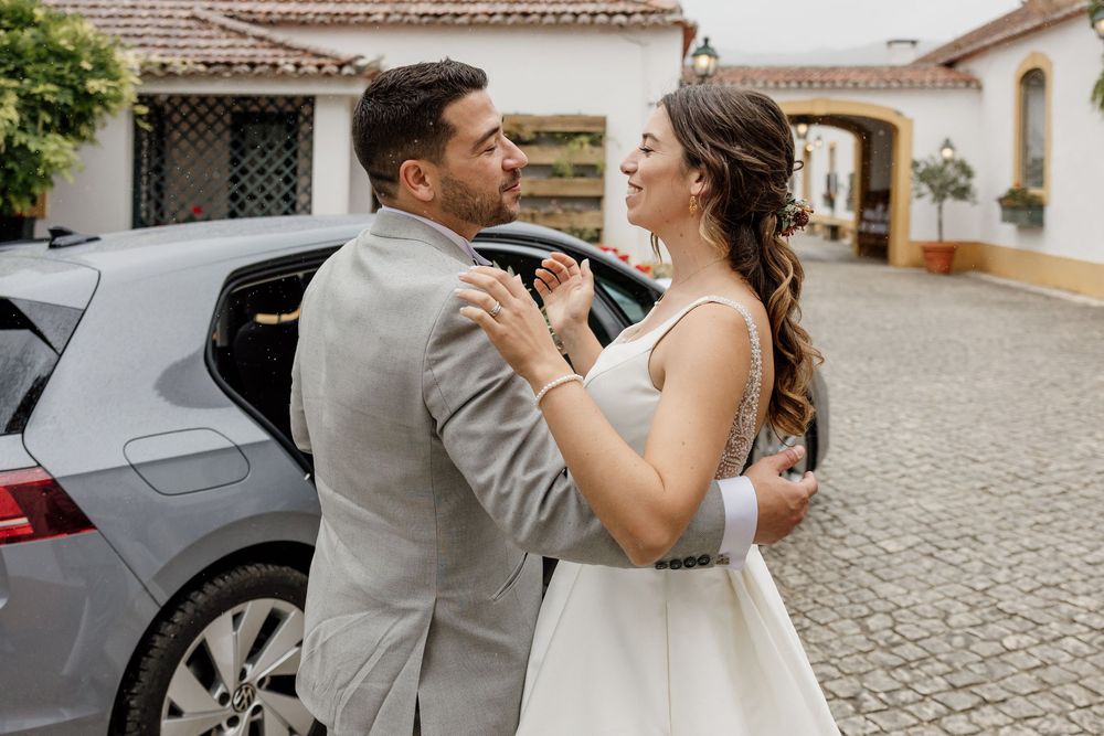 Bride and groom share a romantic moment beside a silver car on cobblestone, dressed in wedding attire.