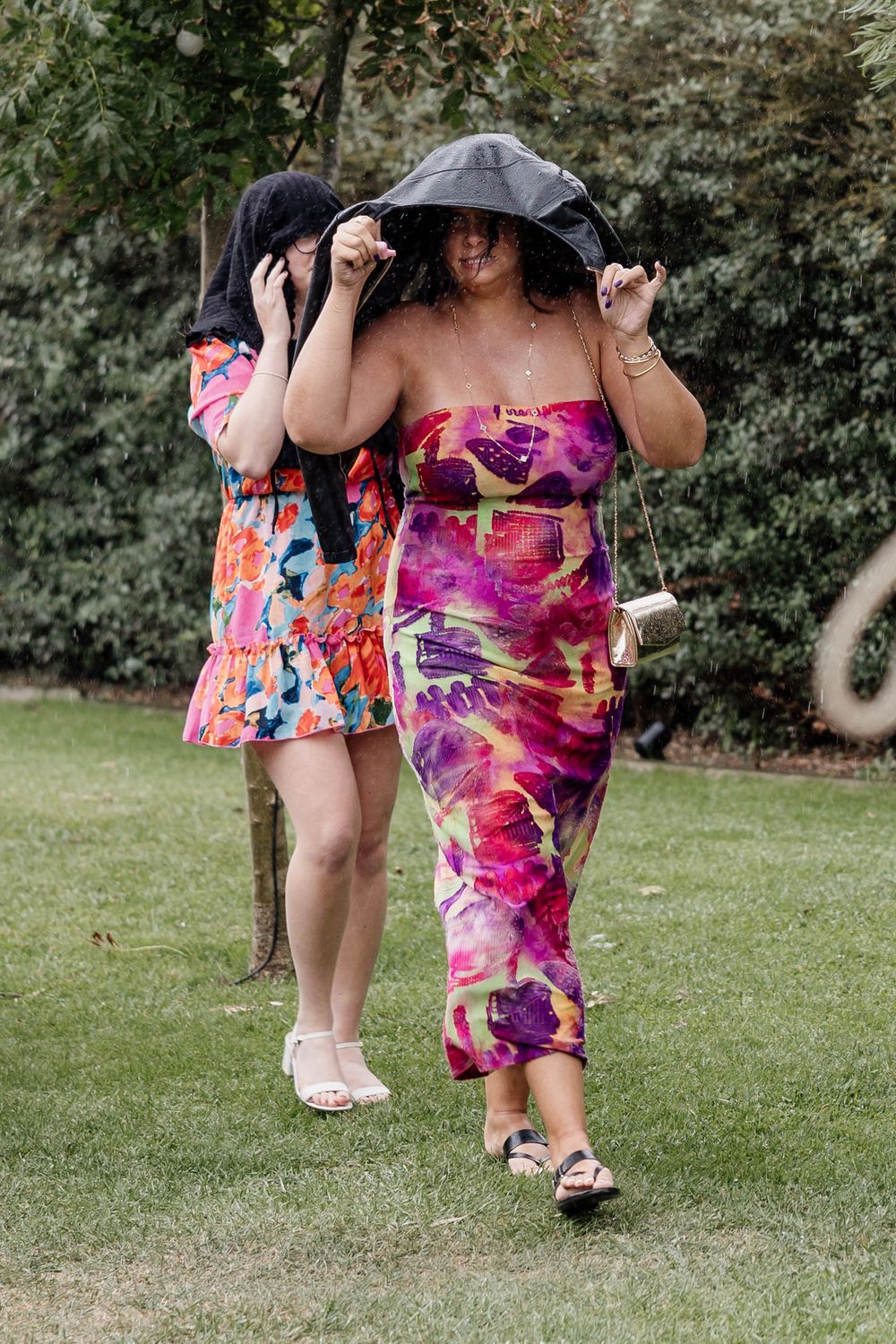 Two women in colorful floral dresses shielding themselves from rain with a black umbrella while walking on grass.