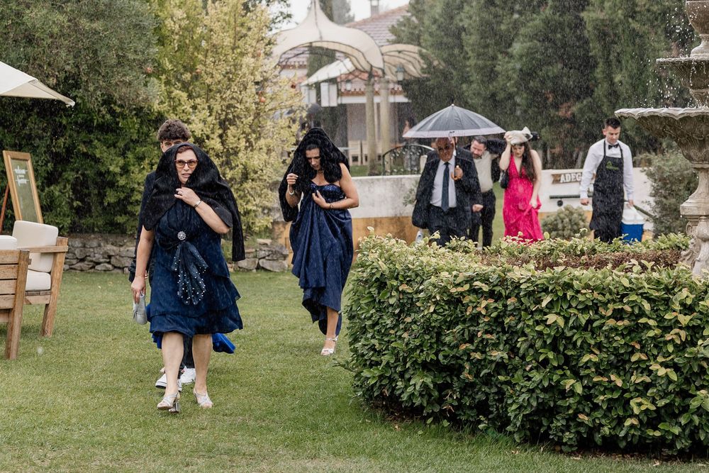 Wedding guests in navy blue dresses walking through garden in light rain, some holding umbrellas at outdoor venue.