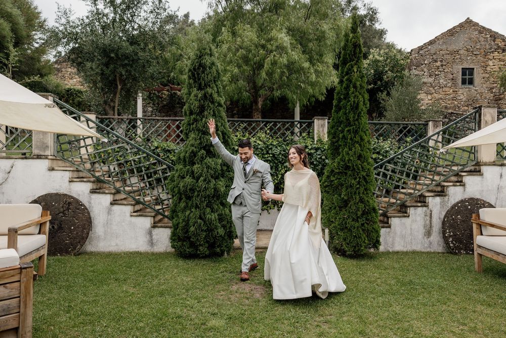 Bride and groom holding hands walking on green lawn with cypress trees and stone staircase at elegant outdoor wedding venue.