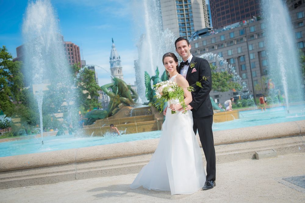 A couple poses for wedding photos in front of a cascading fountain with city buildings in the background.