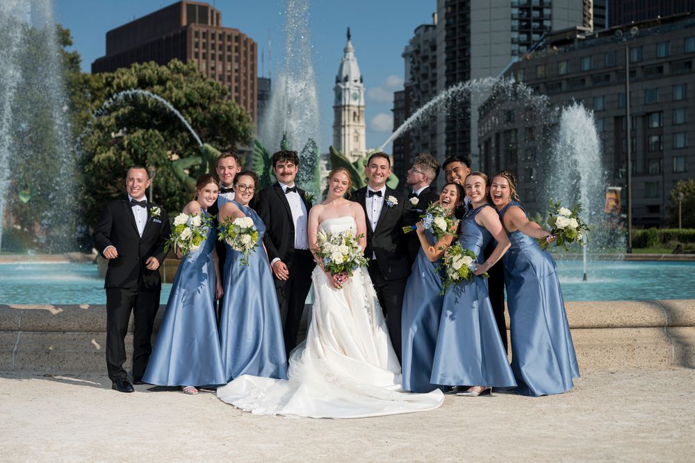 Wedding party in light blue dresses and formal attire poses together in front of city buildings and architecture.