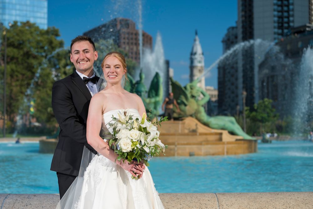 A couple poses for wedding photos in front of Logan Square fountain in Philadelphia on a sunny day.