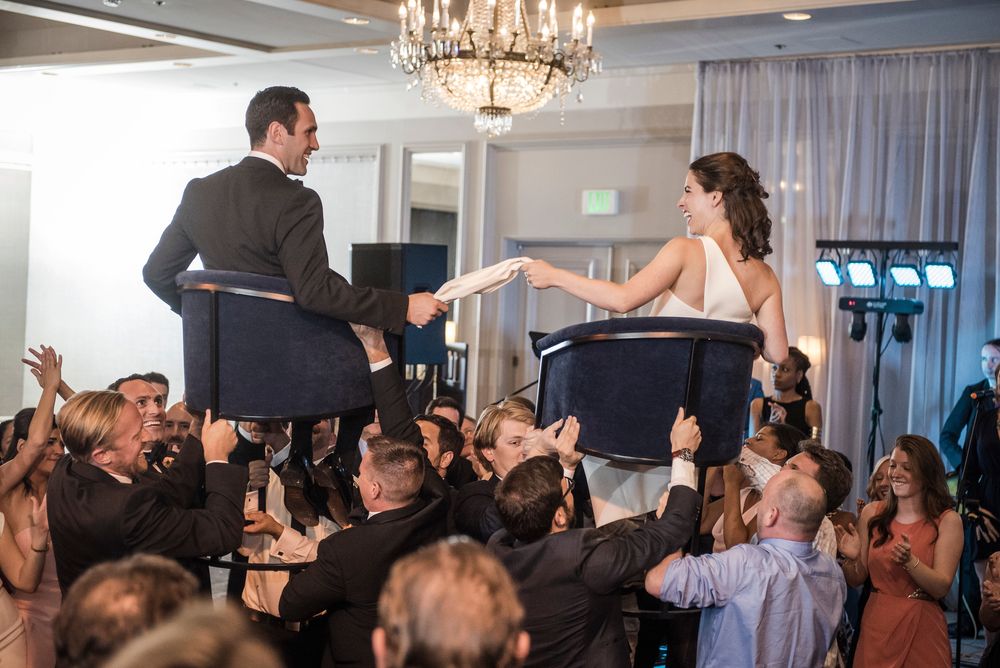 Wedding guests dance and celebrate on a crowded dance floor under elegant crystal chandeliers at an indoor reception.