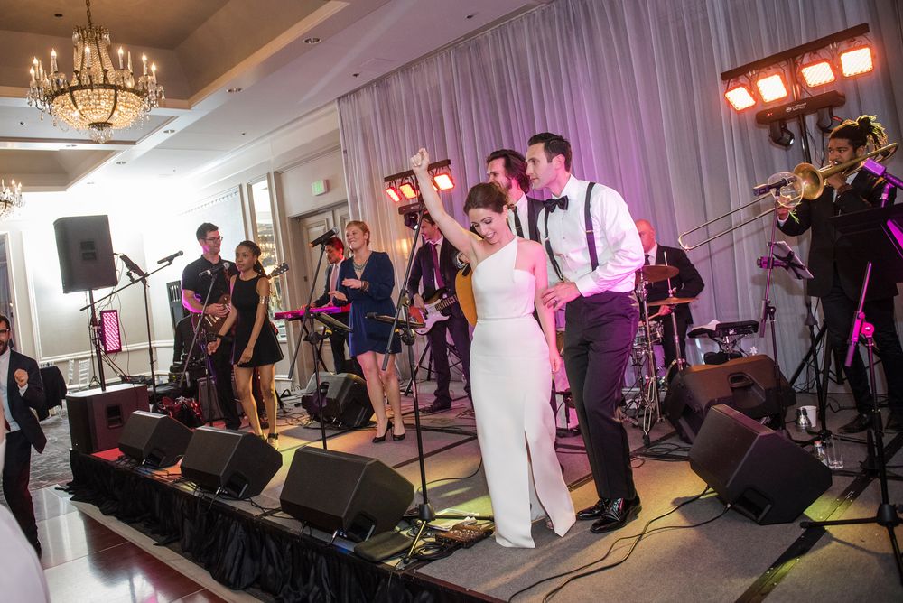 Live band performs on stage at a wedding reception with instruments and pink lighting.