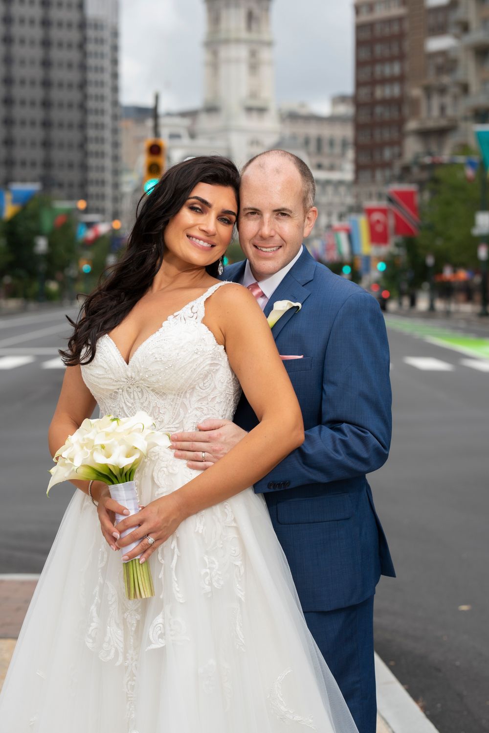 A happy couple poses together on a city street, with the bride in white lace and groom in navy blue suit.