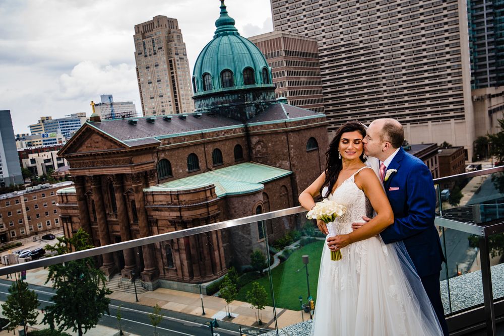 A couple embraces during their wedding photoshoot with a city skyline and church steeple in the background.