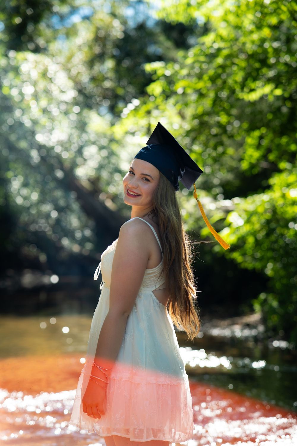 Graduate in a white and pink dress poses by a waterside setting during a sunny graduation photoshoot.