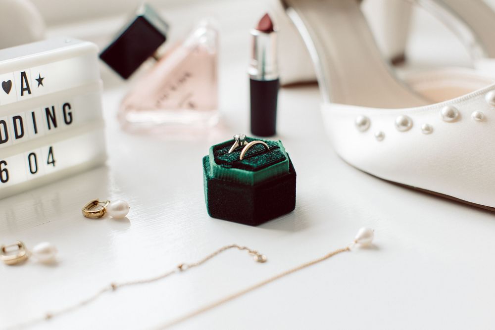 Wedding details featuring white bridal shoes, emerald ring box, and delicate gold jewelry on a light surface.