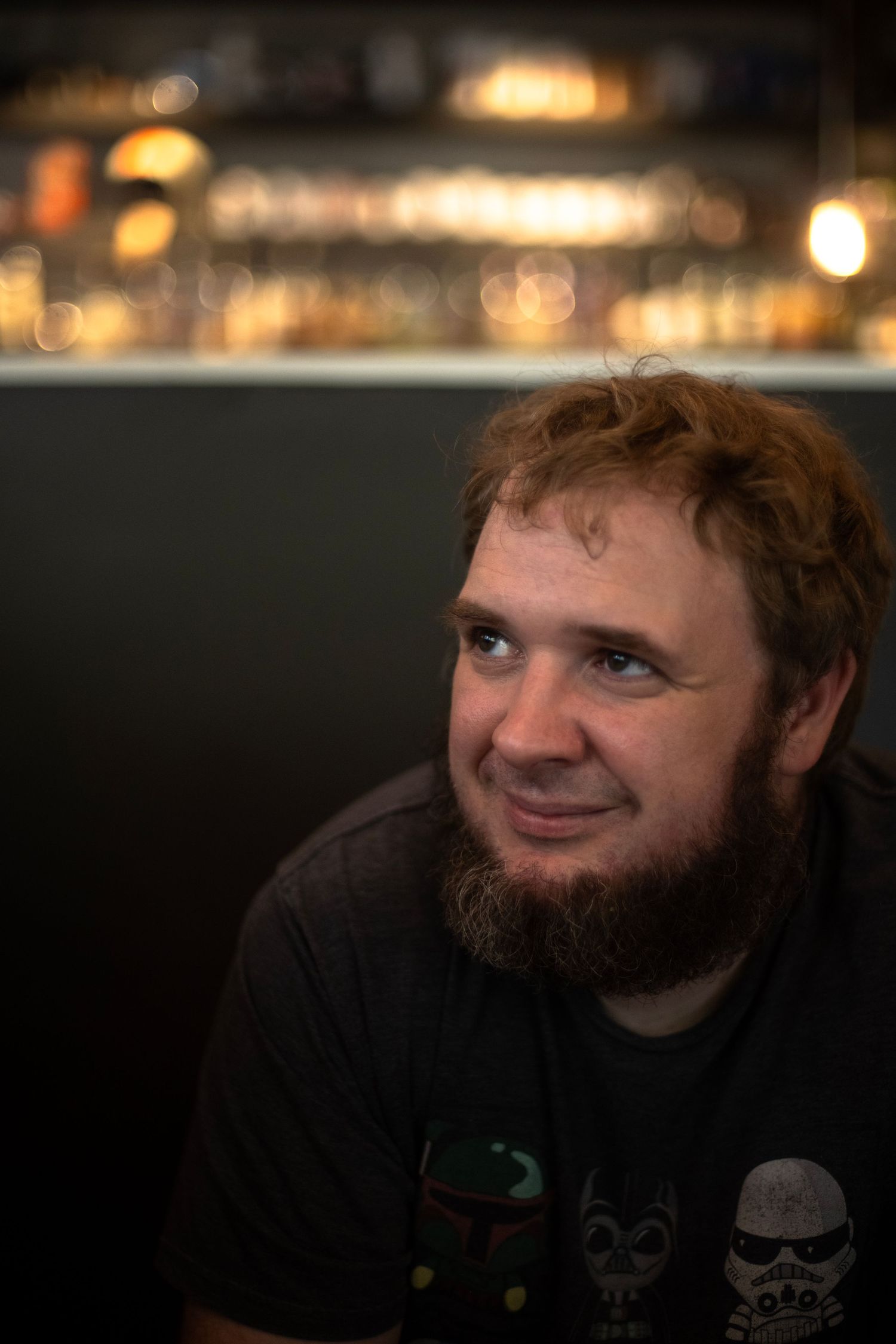 Red-haired bearded man smiling in a dimly lit bar with bokeh bottle lights in the background.