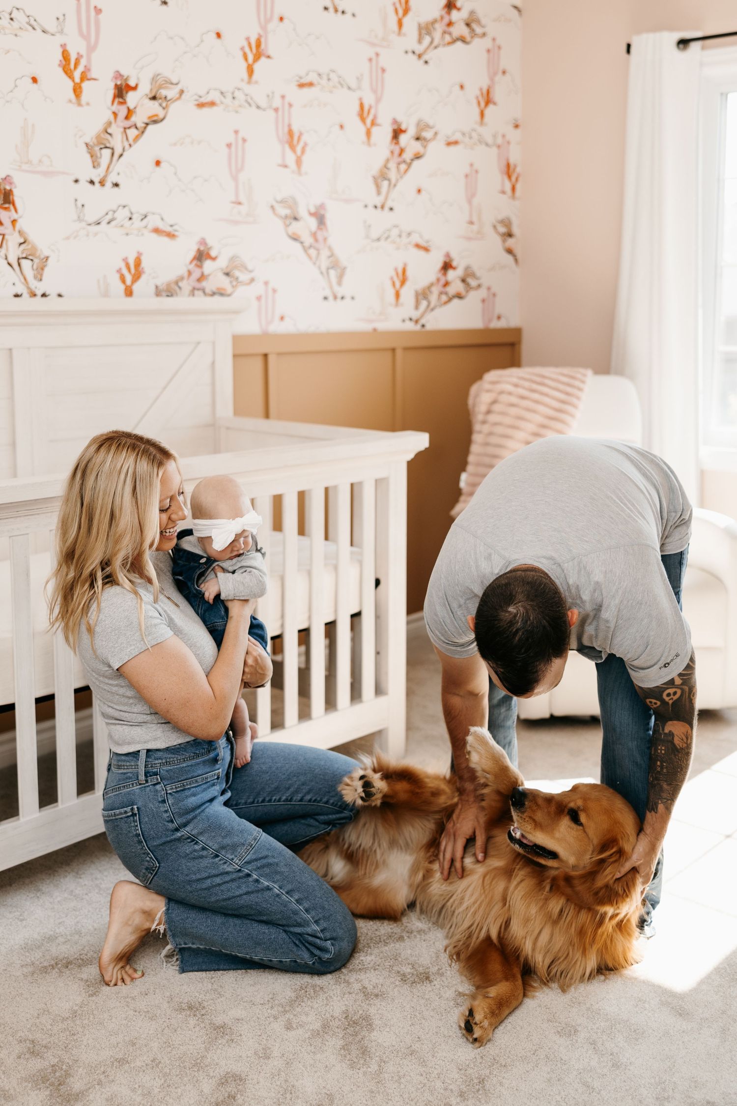 Young family with newborn baby and golden retriever dog in a decorated nursery room with floral wallpaper.