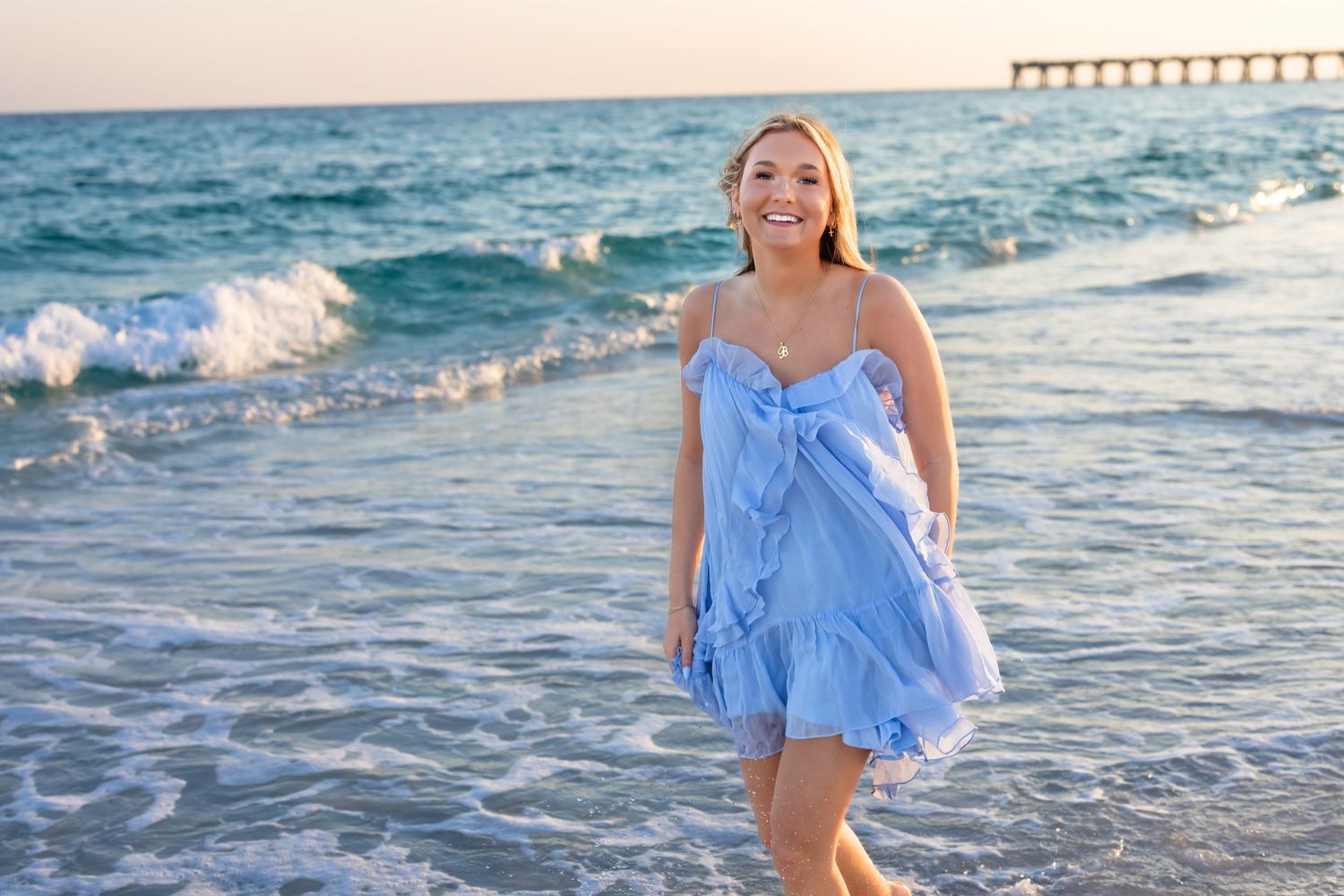 Smiling woman in blue ruffle dress walking on a sunny beach with ocean waves and a pier in the background.