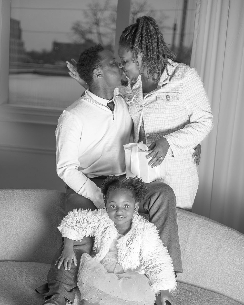 Black and white portrait of a couple sharing a kiss with their young daughter sitting in front.