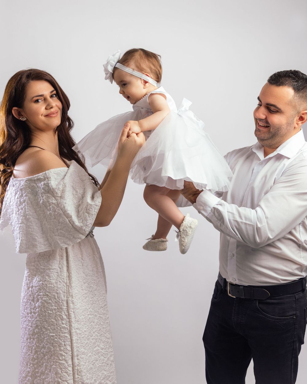 Parents in white outfits lifting their baby girl in a white tutu dress during a family studio portrait.