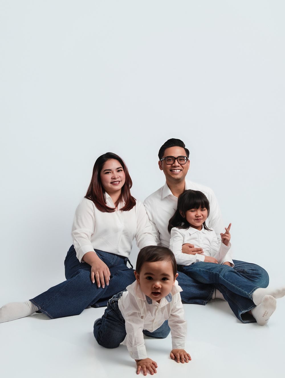 Asian family of four in matching white shirts and jeans posing for a studio portrait with baby crawling in front.