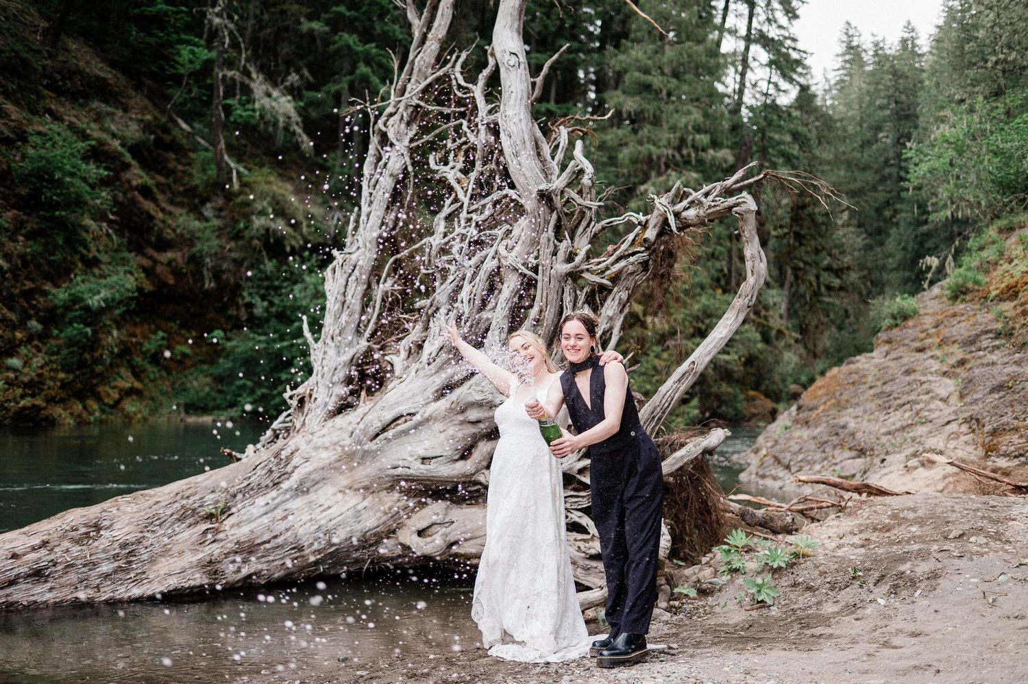 A wedding couple stands on a rocky forest path surrounded by tall evergreen trees while spraying champagne from a bottle