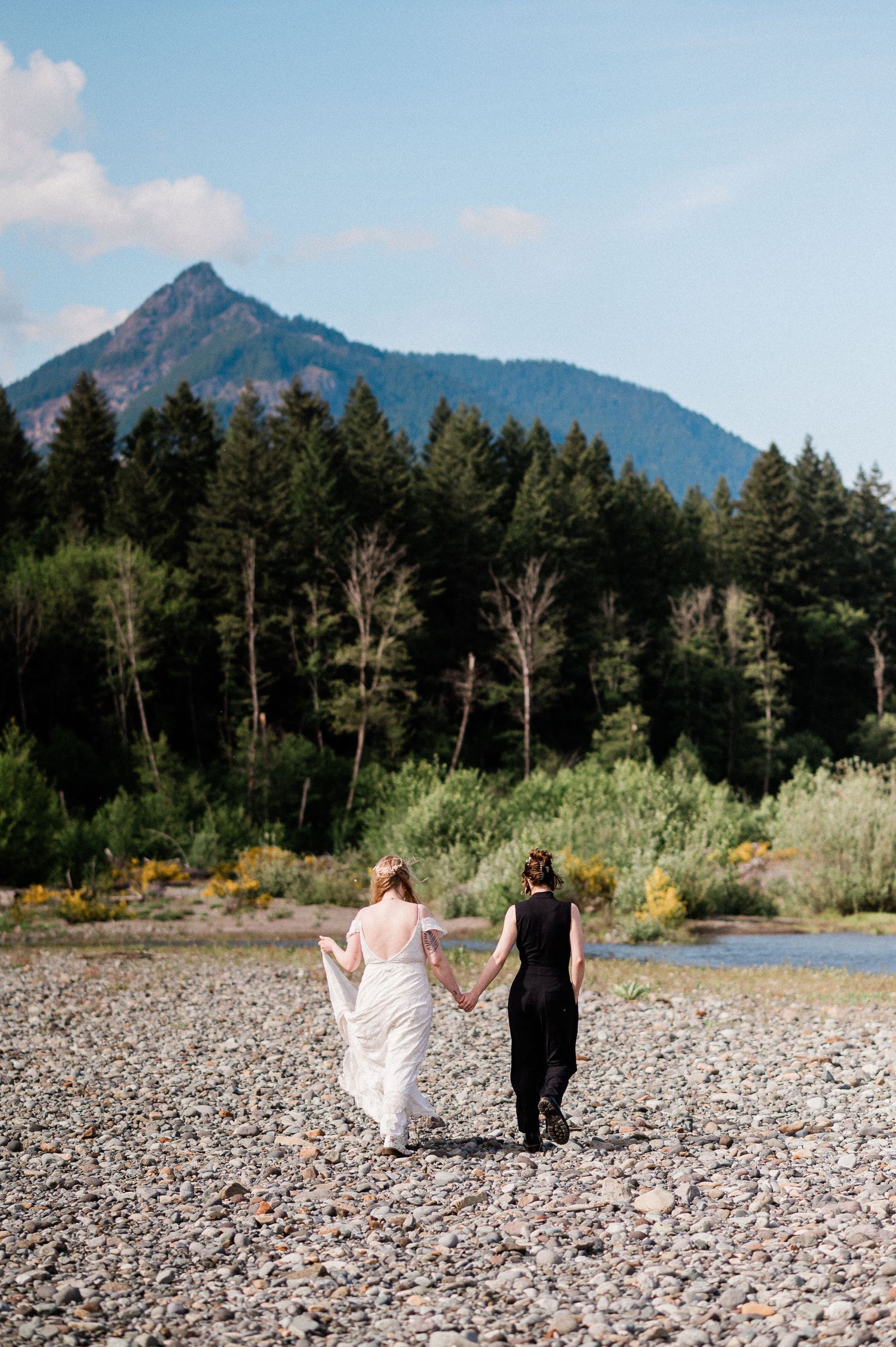 Two people walk along a rocky beach with majestic mountain peaks and evergreen forest in the background.
