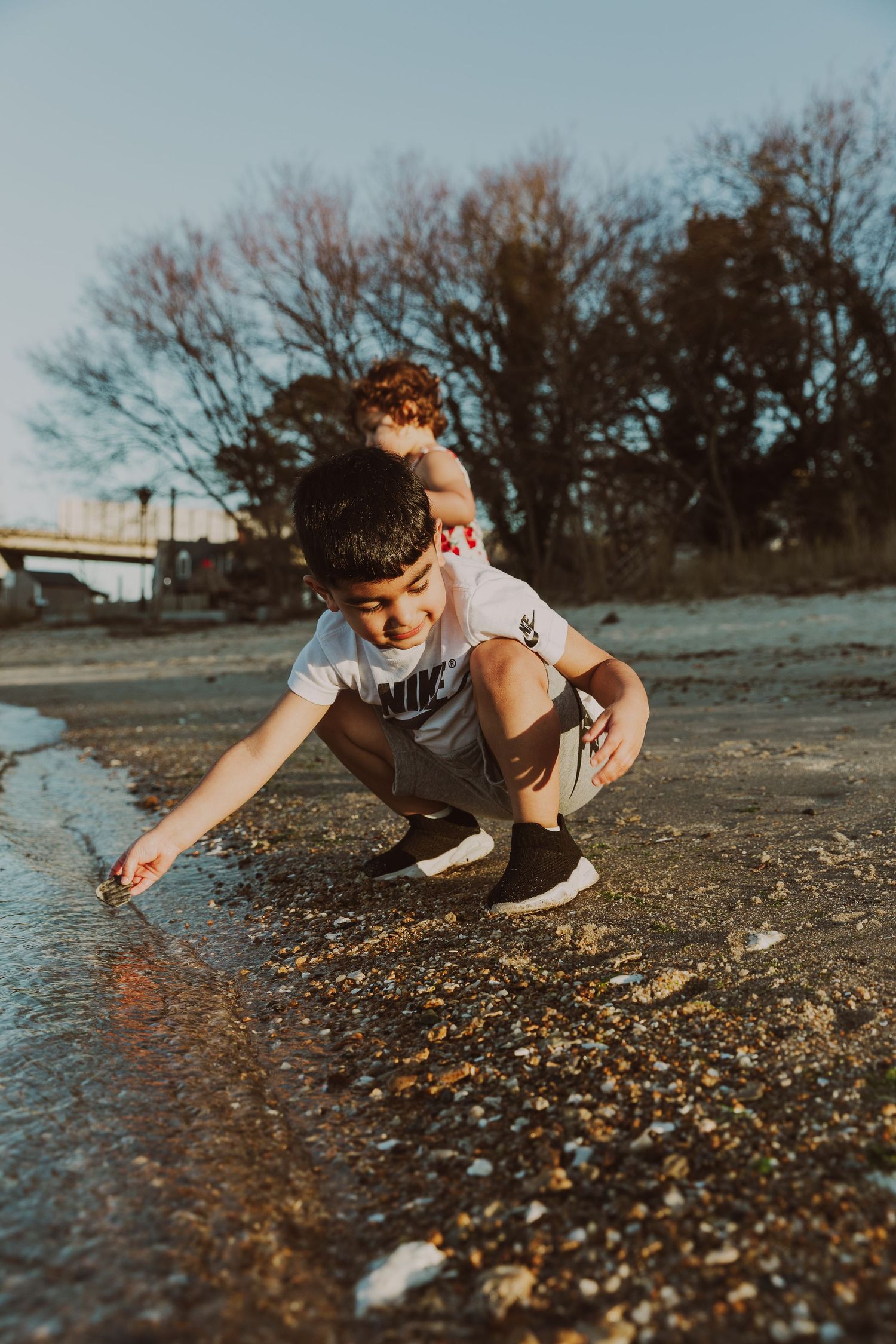 A boy in a Nike shirt crouches by a shallow rocky shoreline at golden hour.