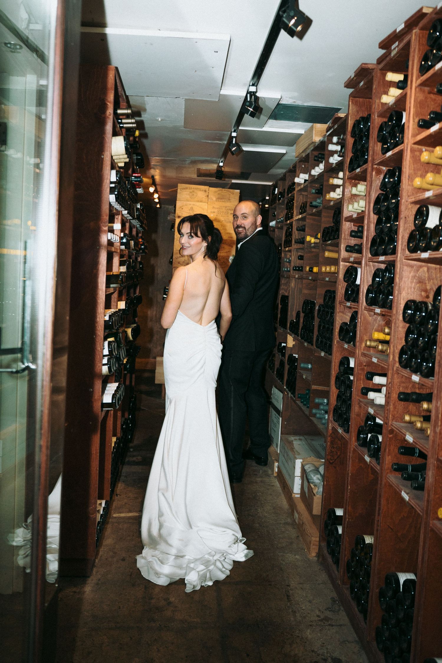 Bride and groom share a romantic moment in a dimly lit wine cellar surrounded by wooden wine racks.