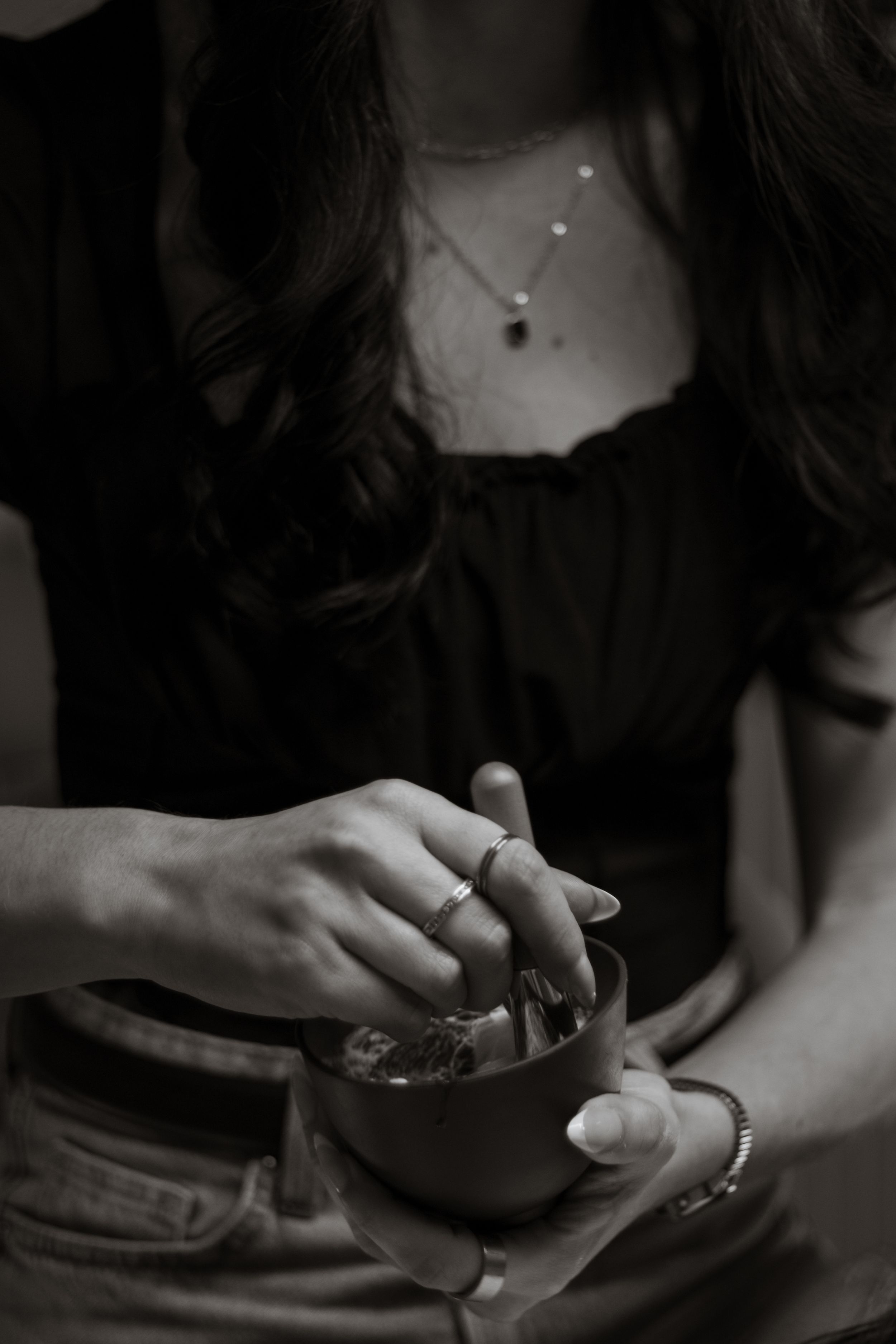 Black and white close-up of woman wearing necklace and ring stirring a small bowl.