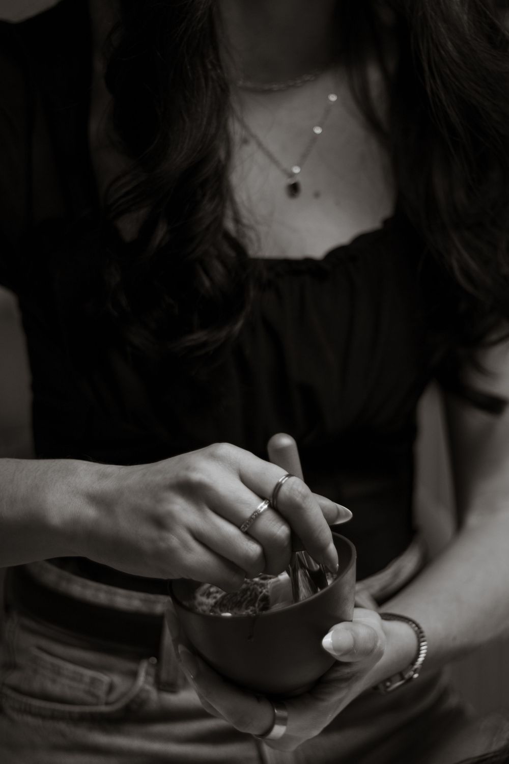 Black and white close-up of woman wearing necklace and ring stirring a small bowl.