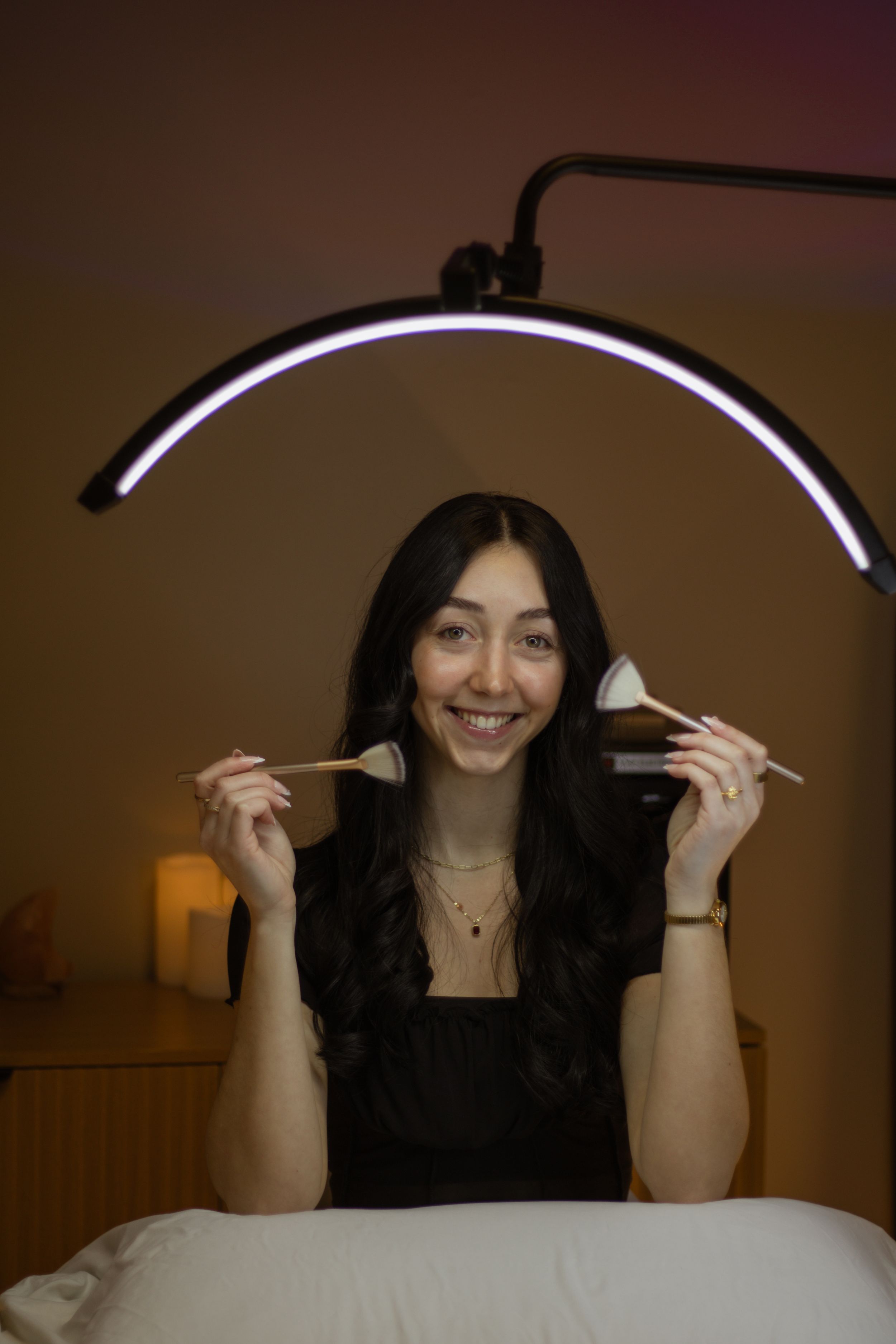 Smiling woman holding makeup brushes under an arc LED ring light in a cozy esthetics studio.